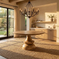 Musa Round Dining Table in a natural wood finish styled in a warm, sunlit dining room with a modern chandelier.